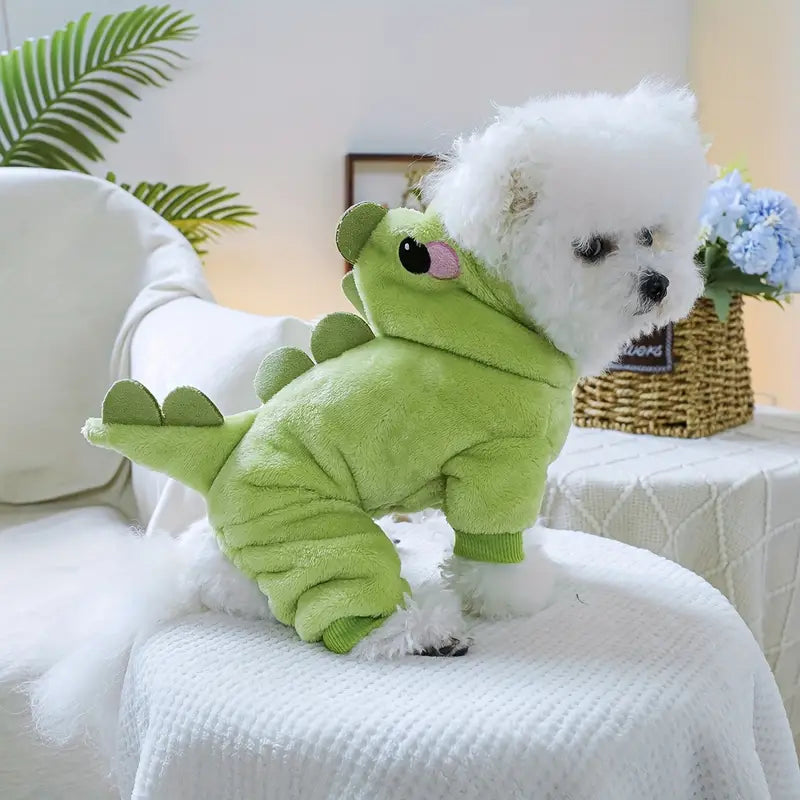 Small white dog wearing a green dinosaur costume on a white couch.