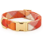 A woven fabric dog collar with a warm orange, red, and cream plaid pattern, featuring a polished gold-tone metal buckle and D-ring, shown on a clean white background.