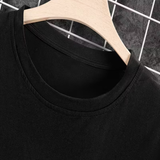 Close-up of a black crew-neck t-shirt hanging on a light wood hanger against a dark gray tiled background with a white wire grid.