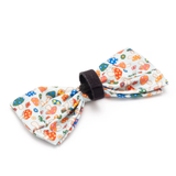 A fabric pet bow tie featuring a colorful mushroom and floral pattern on a white background, secured with a black elastic band.