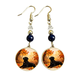 A pair of gold-tone dangle earrings with dark blue beads, clear crystal accents, and round gold-tone cabochons featuring a black Labrador retriever lying in an autumn forest with orange and yellow leaves.