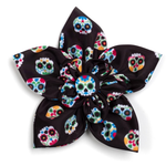 A fabric flower hair clip with a black satin-like base printed with colorful sugar skull motifs, shown on a plain white background.