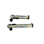 Two fabric pet collars with black bells and black plastic hardware, laid flat on a light whitewashed wood background. The collars have a white base with a colorful pattern of orange and yellow flowers, green leaves, and blue sea plants, and a white label that reads 'PETALS & Purr'.