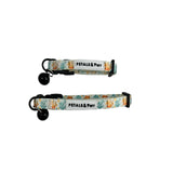 Two fabric pet collars with black bells and black plastic hardware, laid flat on a light whitewashed wood background. The collars have a white base with a colorful pattern of orange and yellow flowers, green leaves, and blue sea plants, and a white label that reads 'PETALS & Purr'.
