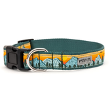 A dog collar with a dark teal-green woven fabric, featuring a printed landscape design of blue mountains and pine trees against an orange and yellow sunset. It has a black plastic buckle and a silver metal D-ring, with a small 'WORTHY OF P&L' brand tag on the side.