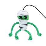 A plush dog toy shaped like a mummy, featuring white and black striped fabric body wraps, a bright green eye band with black stitched eyes and nose, and four green braided rope legs ending in frayed tips. A black hanging loop is attached at the top.