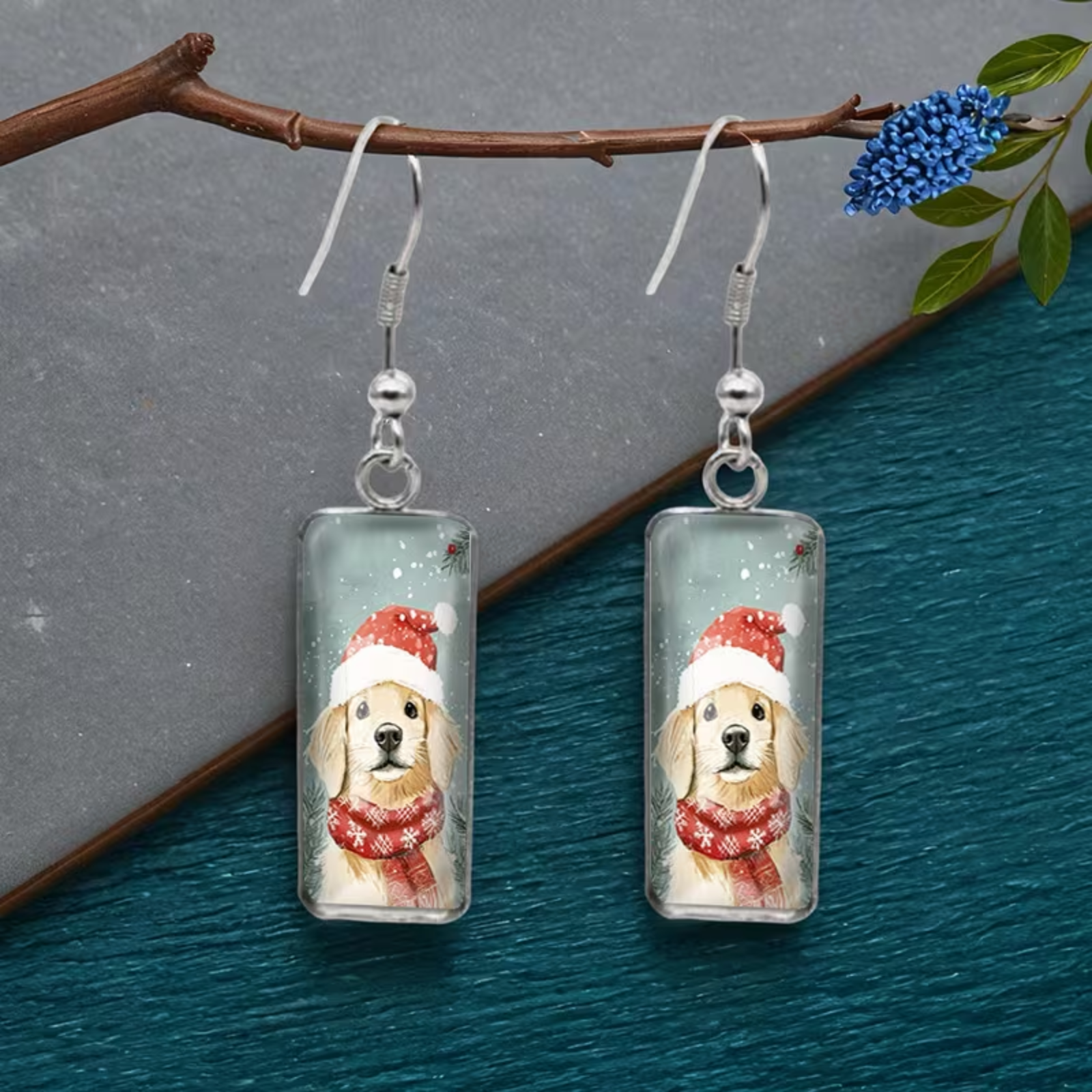 A pair of rectangular drop earrings featuring a printed illustration of a golden retriever dog wearing a red Santa hat and red patterned scarf, suspended from silver fishhook wires. The earrings are displayed on a decorative branch over a split gray and teal textured background.