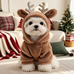 A small white dog wearing a brown plush reindeer costume with antlers, sitting on a carpeted floor in a cozy living room with a sofa, pillows, and a decorated Christmas tree in the background.