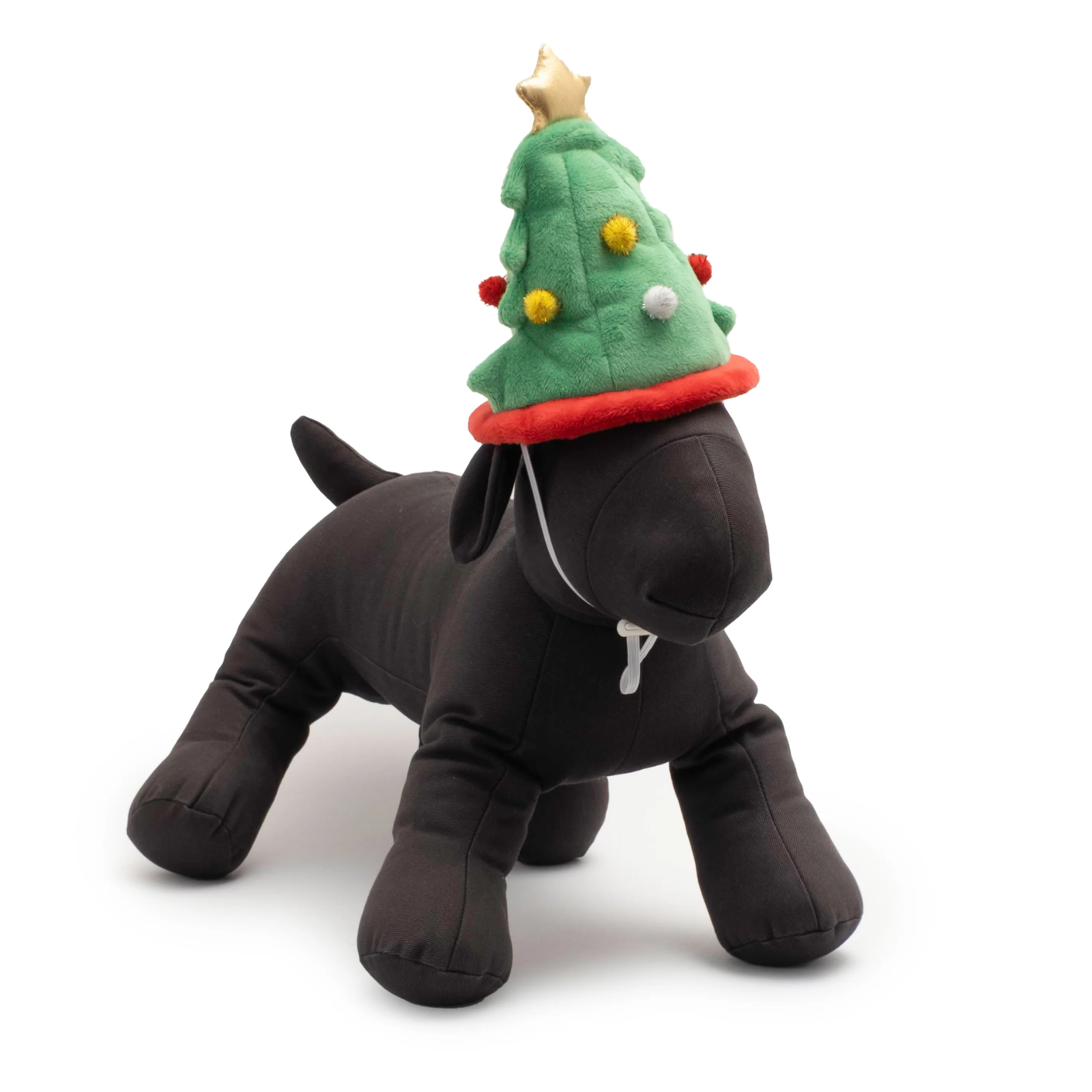 A black plush dog toy wearing a green Christmas tree hat with red trim, yellow and white pom-poms, and a gold star on top, photographed on a clean white background.