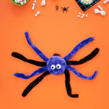 A plush spider toy with a blue-purple round body, white eyes, and black legs, laid flat on an orange background with Halloween-themed props around the edges.