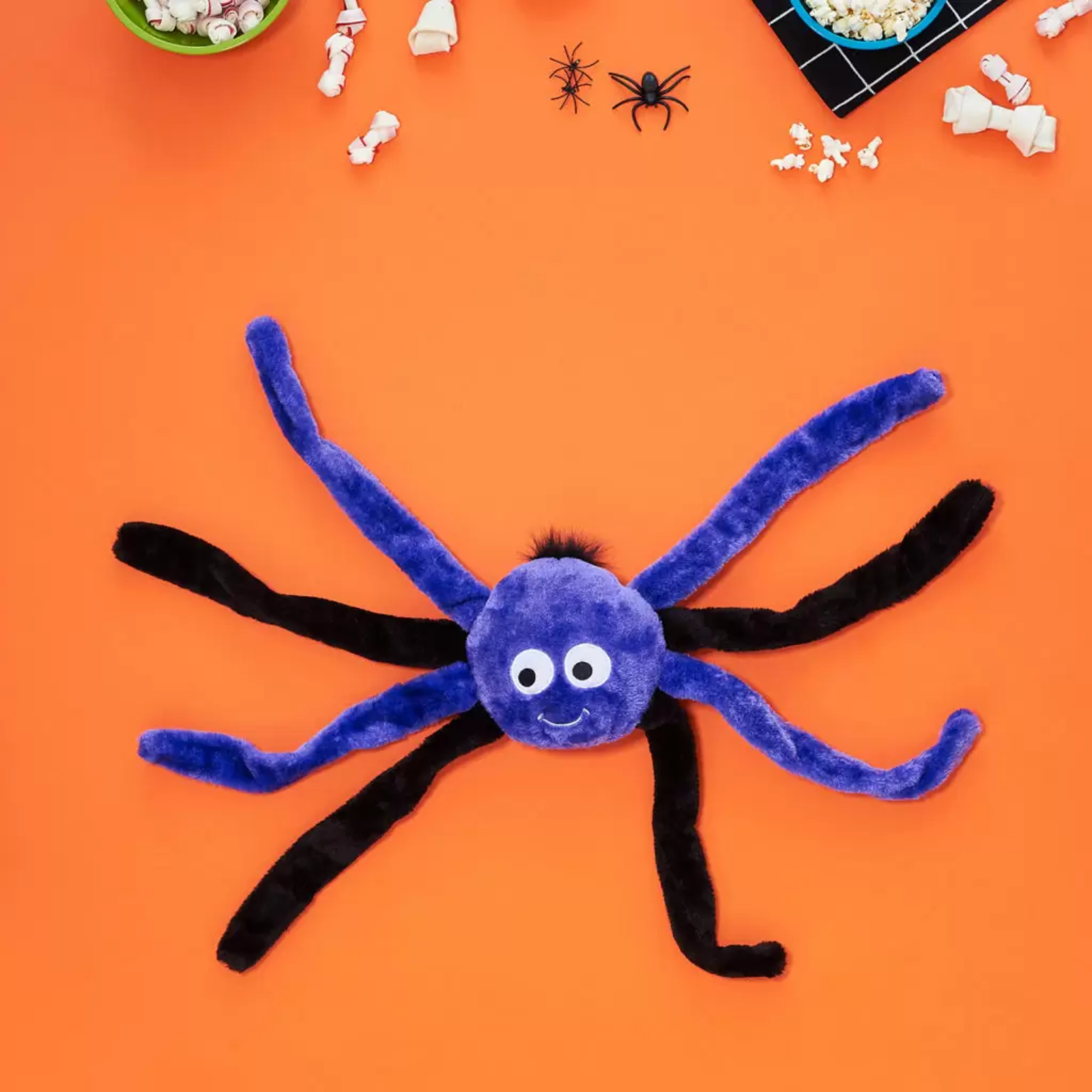 A plush spider toy with a blue-purple round body, white eyes, and black legs, laid flat on an orange background with Halloween-themed props around the edges.