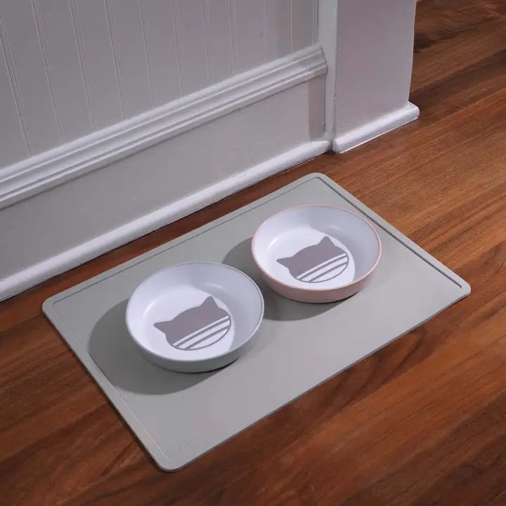 Two pet bowls with cat icons on a mat on a wooden floor.