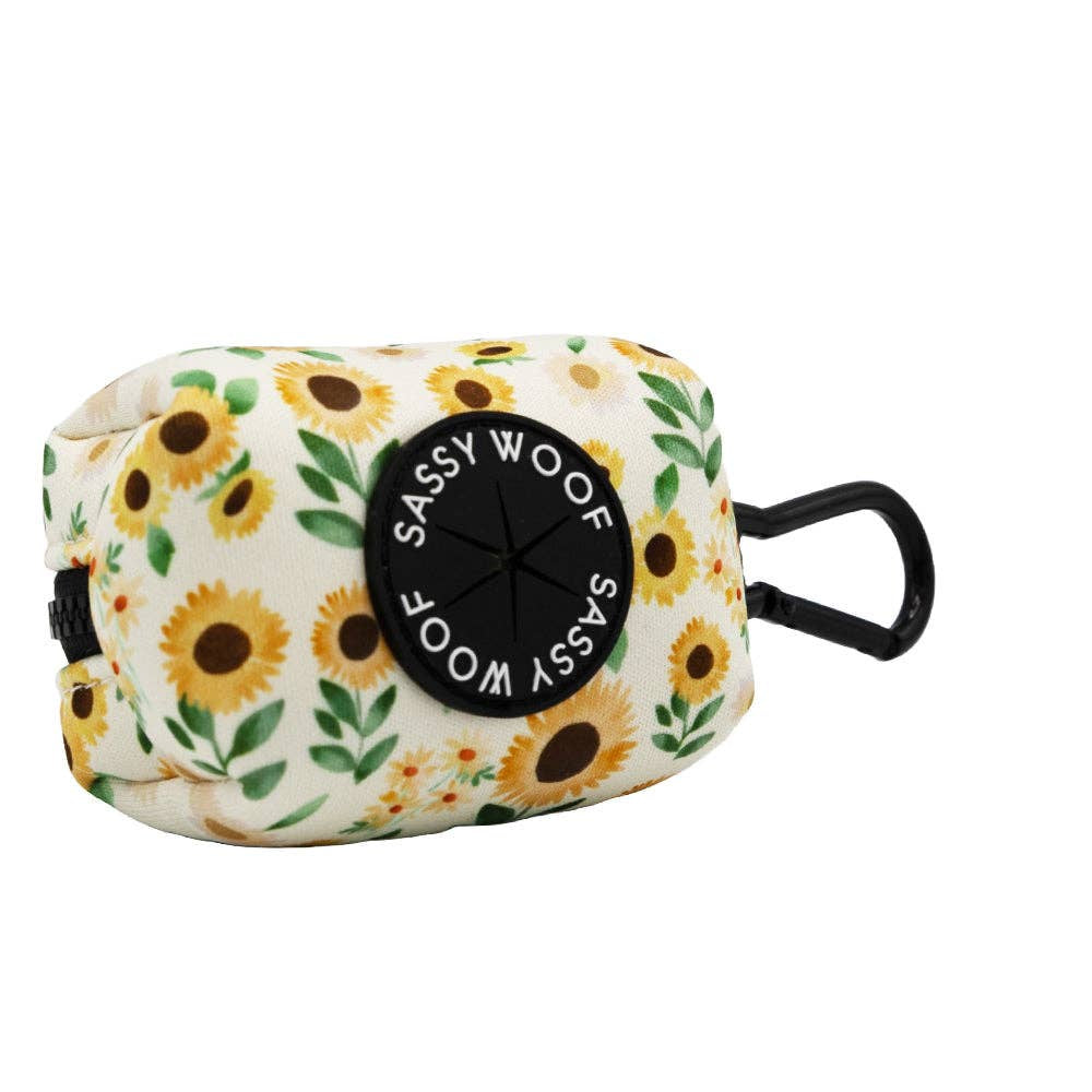 A Sassy Woof dog waste bag pouch featuring a sunflower and green leaf pattern on a cream background, with a black circular logo patch and a black hanging loop.