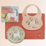 Children's dining set with bib, plate, and spoon featuring unicorn design on a pink background.