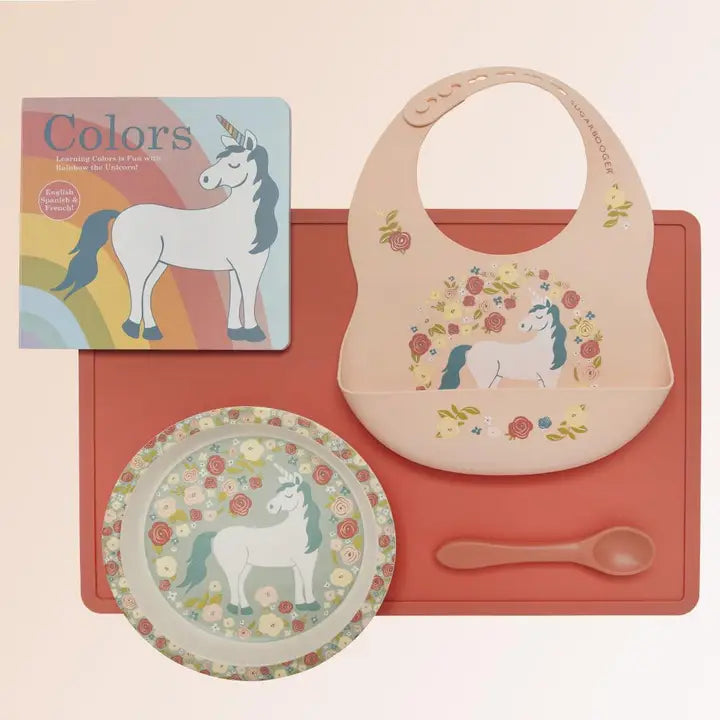 Children's dining set with bib, plate, and spoon featuring unicorn design on a pink background.