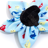 A fabric flower-shaped hair accessory with light blue petals printed with cartoon whales in red, blue, and yellow, secured with a black velcro center on a white background.