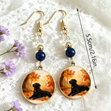 A pair of gold-tone dangle earrings with dark blue beads and oval charms featuring a black dog in an autumn scene, displayed on a white lace background with a 5cm/2.16in size marker.