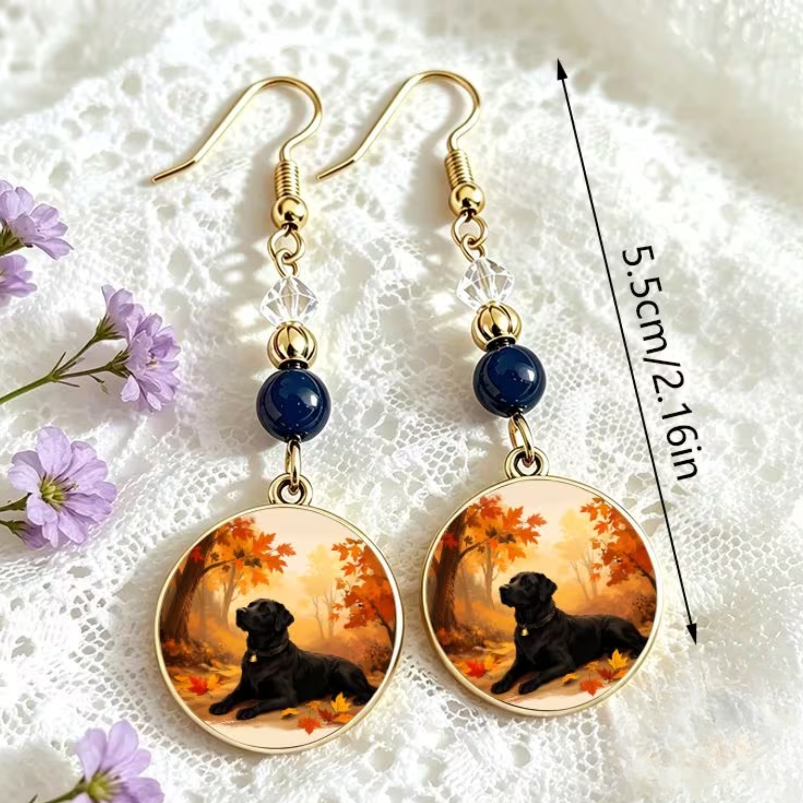 A pair of gold-tone dangle earrings with dark blue beads and oval charms featuring a black dog in an autumn scene, displayed on a white lace background with a 5cm/2.16in size marker.