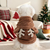 A small white dog seen from behind wearing a brown plush pet sweater with antler details on the back, sitting in a cozy living room with a Christmas tree and holiday decor in the background.