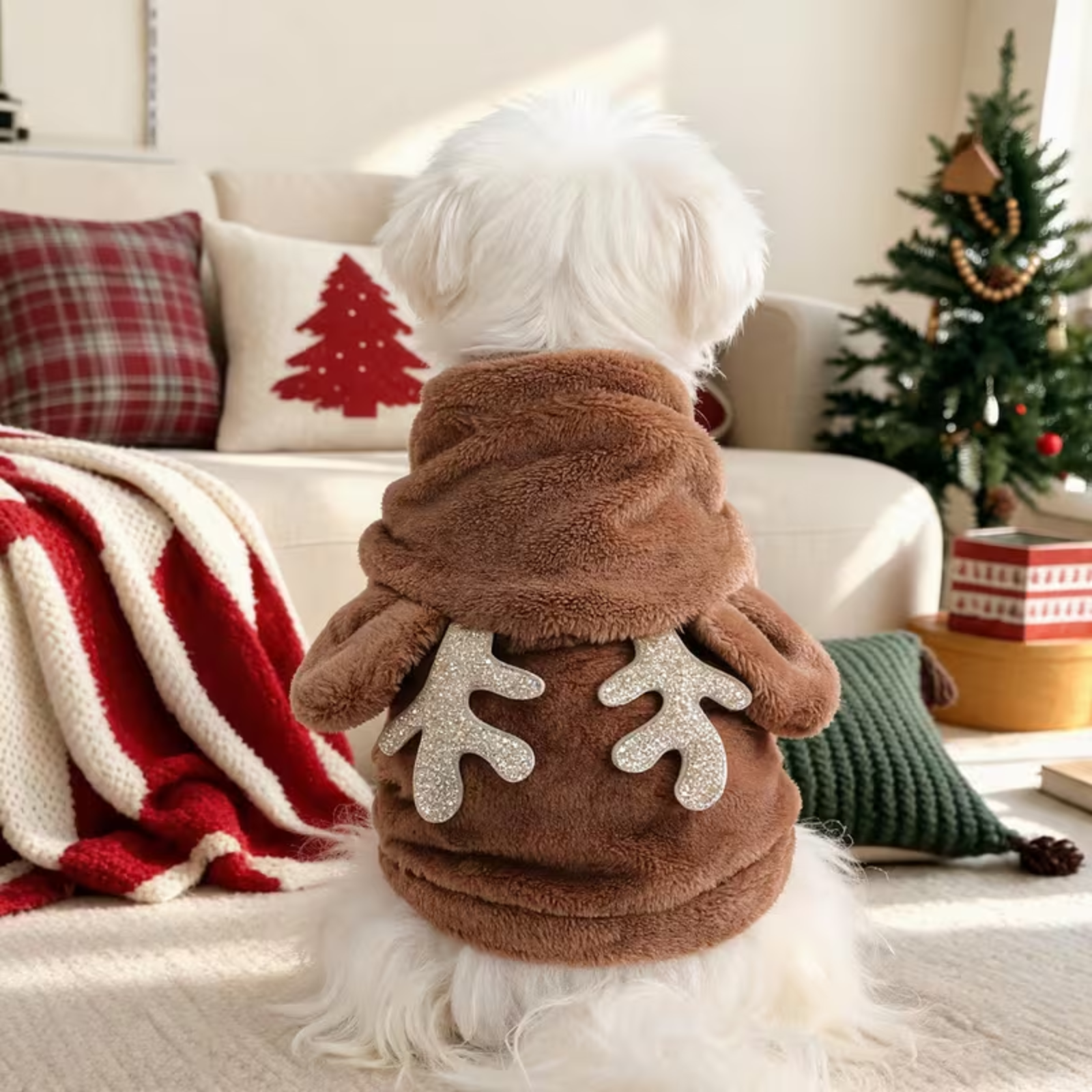 A small white dog seen from behind wearing a brown plush pet sweater with antler details on the back, sitting in a cozy living room with a Christmas tree and holiday decor in the background.