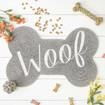Gray bone-shaped mat with 'Woof' text on a white wooden surface with dog toys and treats.
