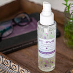Bottle of Pet House room spray on a wooden surface with a laptop and glasses in the background.