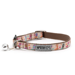A patterned cat collar with a silver bell and a gray metal nameplate that reads 'WORTHY CAT'. The collar features a whimsical multicolor print of boots, stars, and paw prints on a light beige background, with a solid brown inner lining.