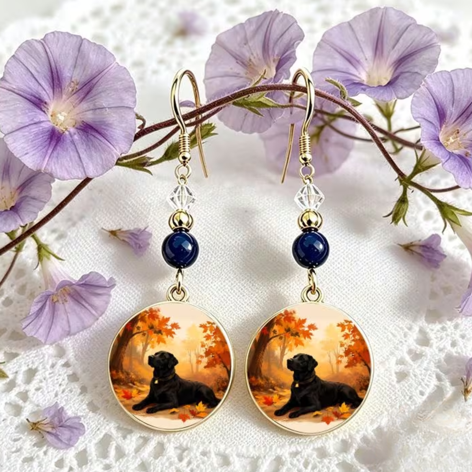A pair of gold-tone dangle earrings with dark blue beads and clear faceted beads, featuring round glass charms printed with a black dog lying in an autumn forest. The earrings are displayed on a white lace surface with purple flowers in the background.