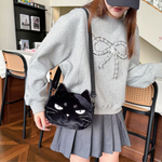A person wearing a gray sweatshirt and pleated gray skirt carries a black plush cat-face crossbody bag with white eyes and a bow-shaped strap in a home interior setting.
