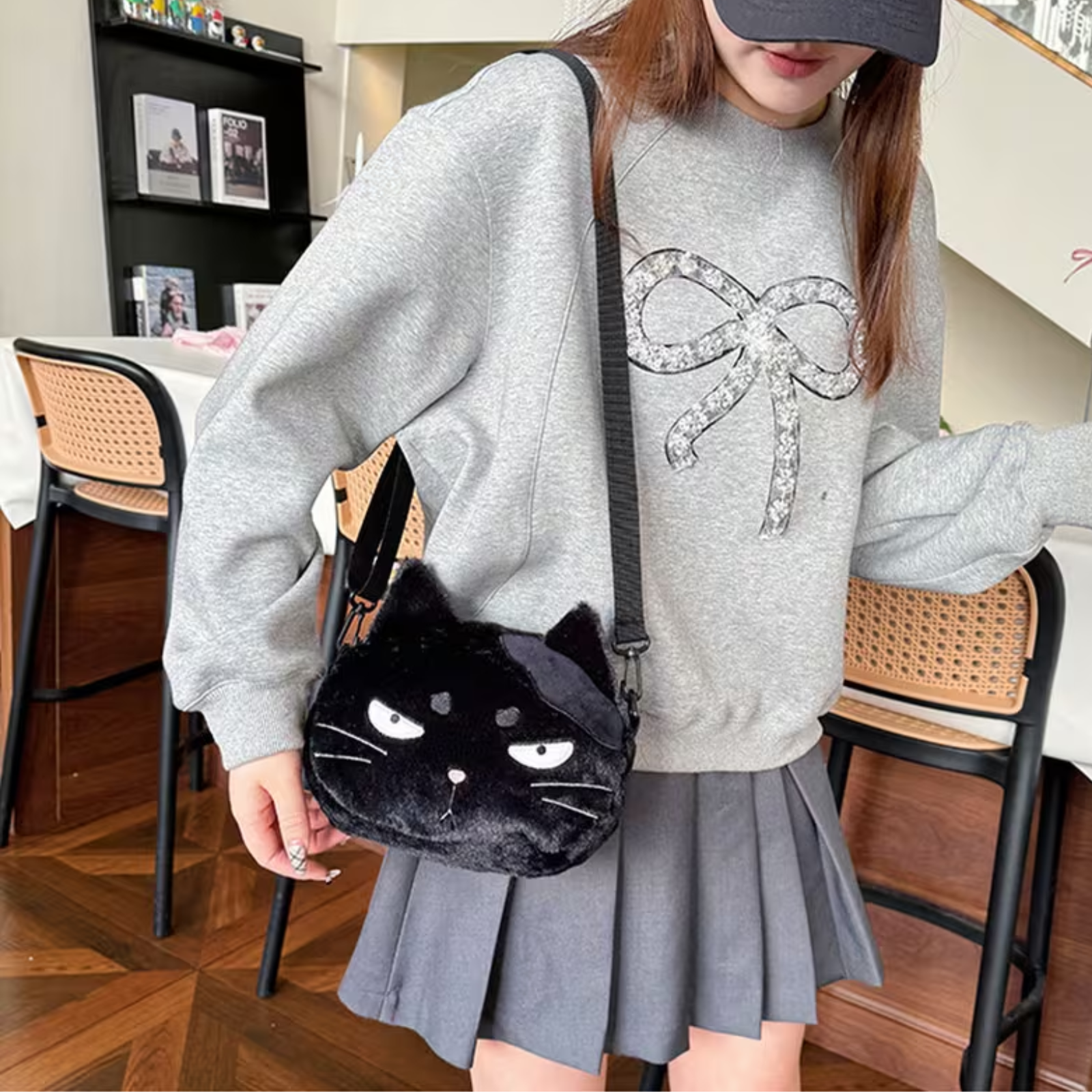A person wearing a gray sweatshirt and pleated gray skirt carries a black plush cat-face crossbody bag with white eyes and a bow-shaped strap in a home interior setting.