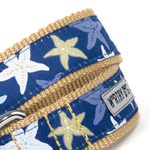 Close-up of a woven dog collar with a navy blue fabric featuring white, light blue, and yellow starfish patterns, tan webbing, and a metal buckle. A fabric label reads 'WORTH DOG'.