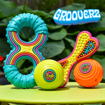 A set of colorful dog toys including a turquoise ring teether with rainbow stripes, a yellow heart-shaped toy with pink trim, a lime green ball, and an orange ball, displayed on a green surface with a leafy background. The brand name 'GROOVERZ' is displayed in a blue 3D font at the top right.