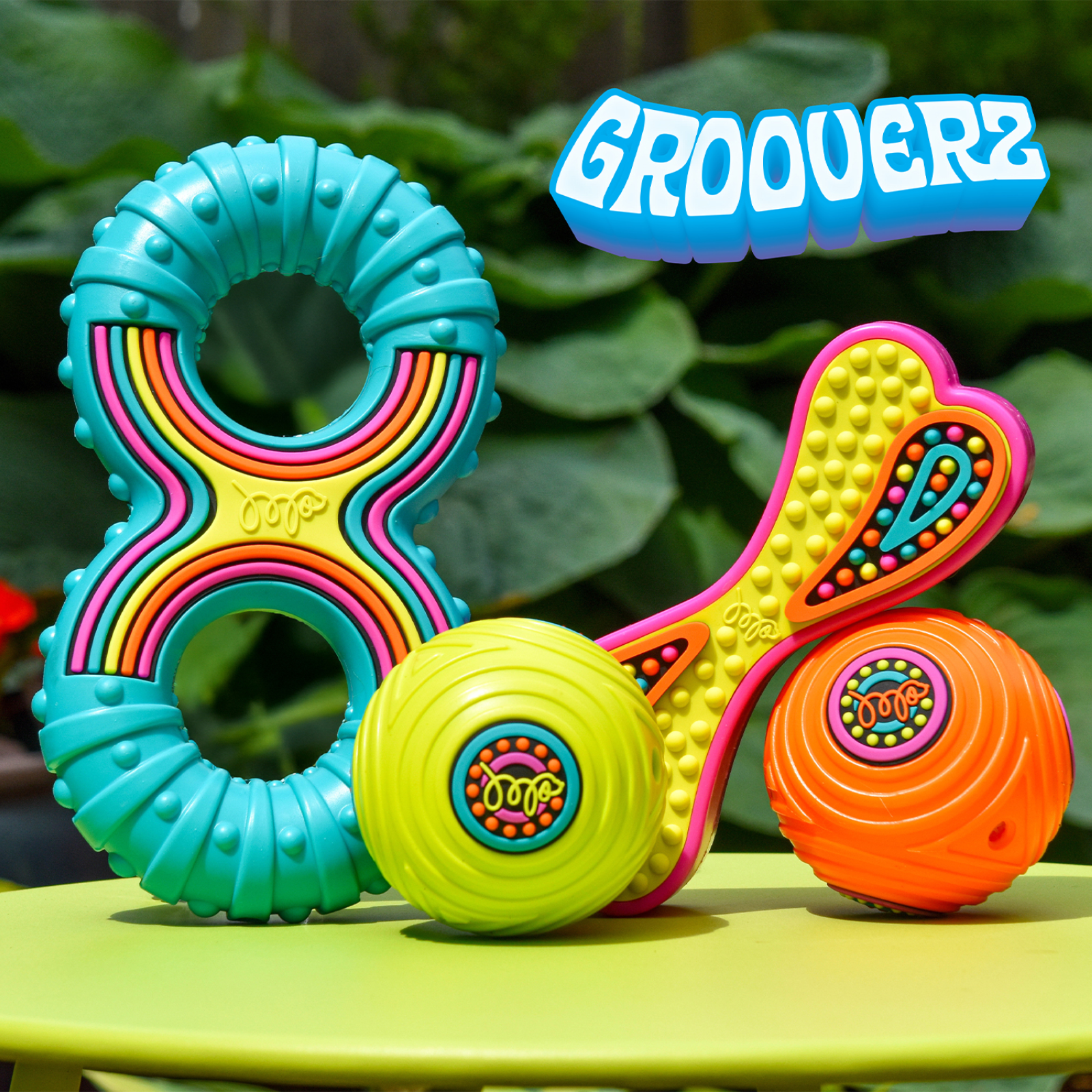 A set of colorful dog toys including a turquoise ring teether with rainbow stripes, a yellow heart-shaped toy with pink trim, a lime green ball, and an orange ball, displayed on a green surface with a leafy background. The brand name 'GROOVERZ' is displayed in a blue 3D font at the top right.