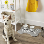 Dog sitting on a wooden floor with two pet bowls on a decorative mat that says 'Woof'.