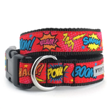 A red dog collar with a black woven trim and a black plastic buckle, featuring a colorful comic book style print with text like 'BOOM!', 'BLAM!', 'SMASH!', 'POW!', and 'HAC!' in yellow, blue, and orange. A silver metal ring is attached on the side.