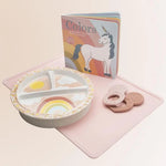 Children's plate with rainbow design, coloring book with unicorn, and cookies on a pink surface
