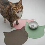 Cat standing on a pink cat-shaped mat with two other mats and a small bowl on a white background