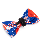 A patriotic pet bow tie featuring a red, white, and blue American flag pattern with stars, stripes, and chevron designs, secured with a black velcro strap on a white background.