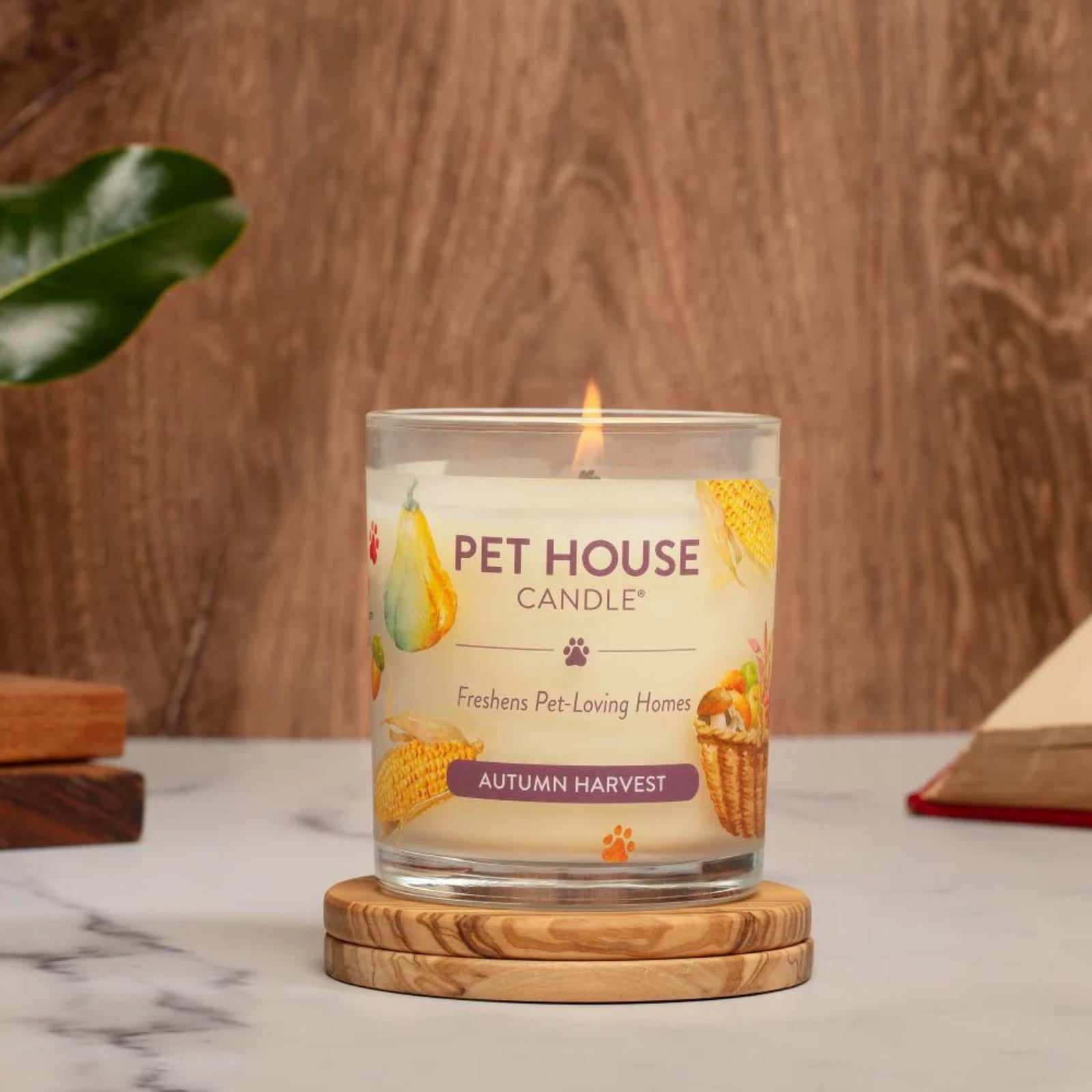 Pet House Candle labeled 'Autumn Harvest' on a wooden coaster with a floral and book background.