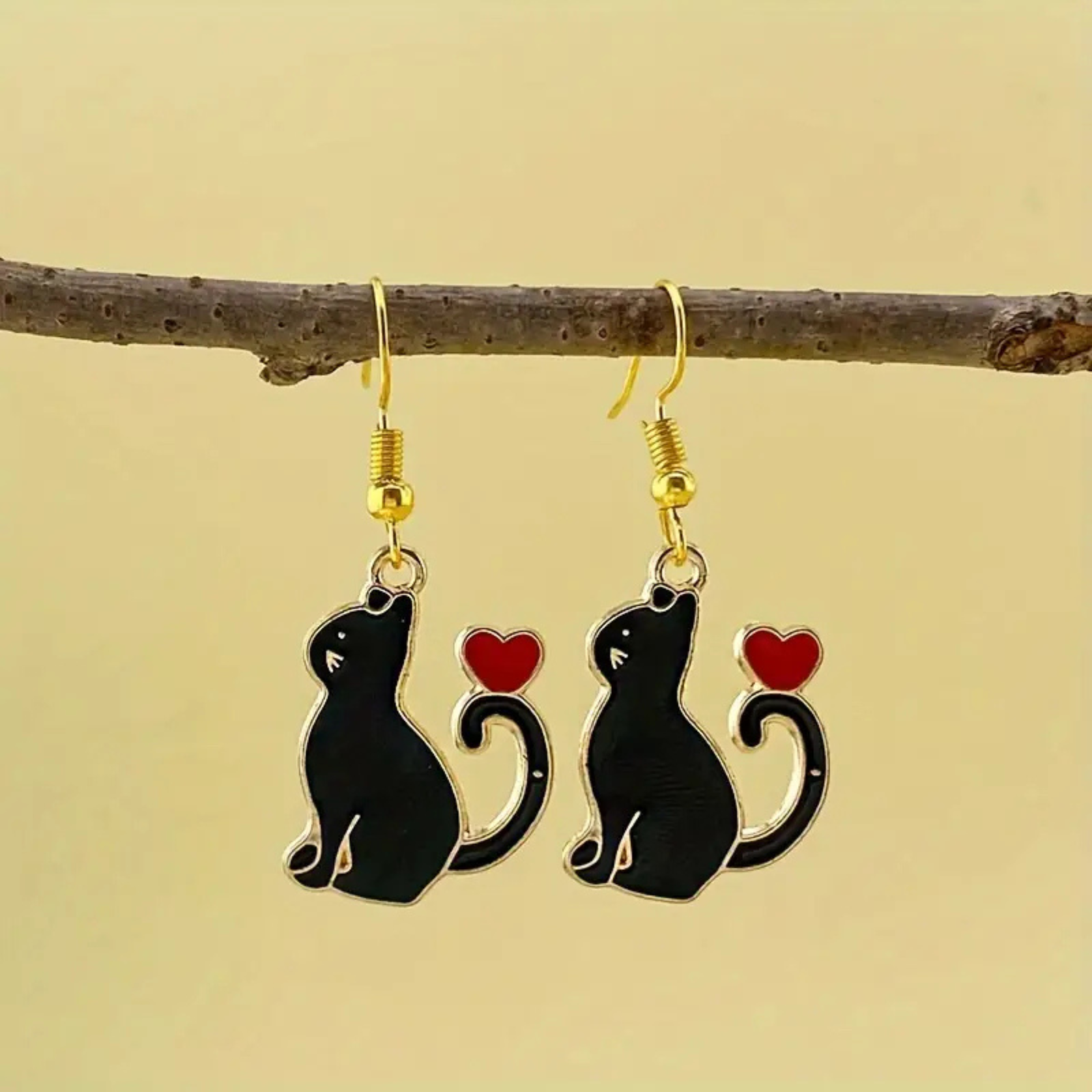 Black cat earrings with red hearts hanging from a branch on a beige background