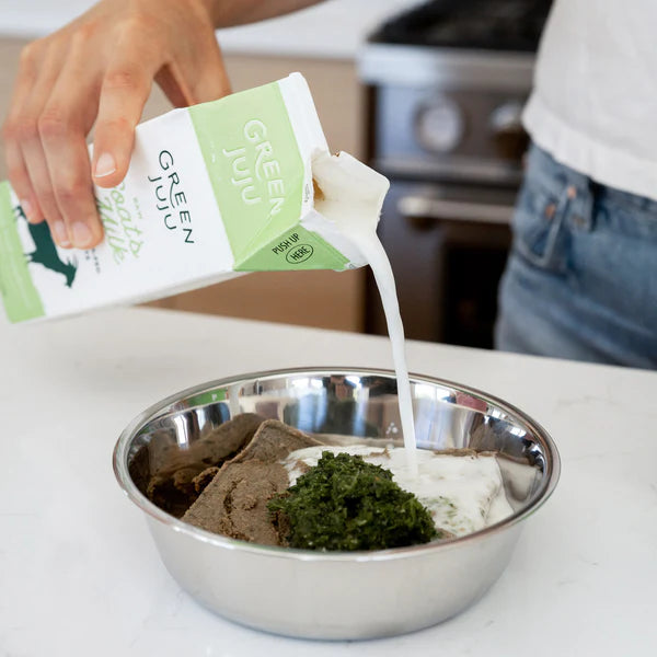 Person pouring milk from a Green Juju carton into a metal bowl with pet food.