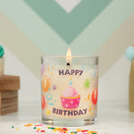 Glass candle with 'Happy Birthday' text and cupcake design, lit on a decorative background.