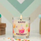 Glass candle with 'Happy Birthday' text and cupcake design, lit on a decorative background.