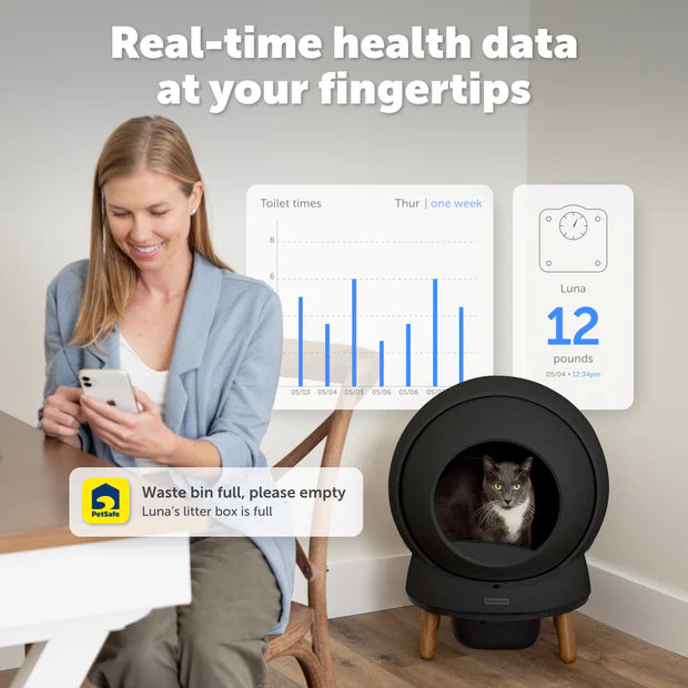 Woman using a smartphone with a smart pet litter box and monitoring app on a screen.