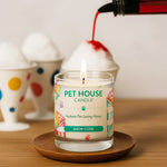 Pet House Candle labeled 'Snow Cone' being lit on a wooden surface with colorful ceramic items in the background.