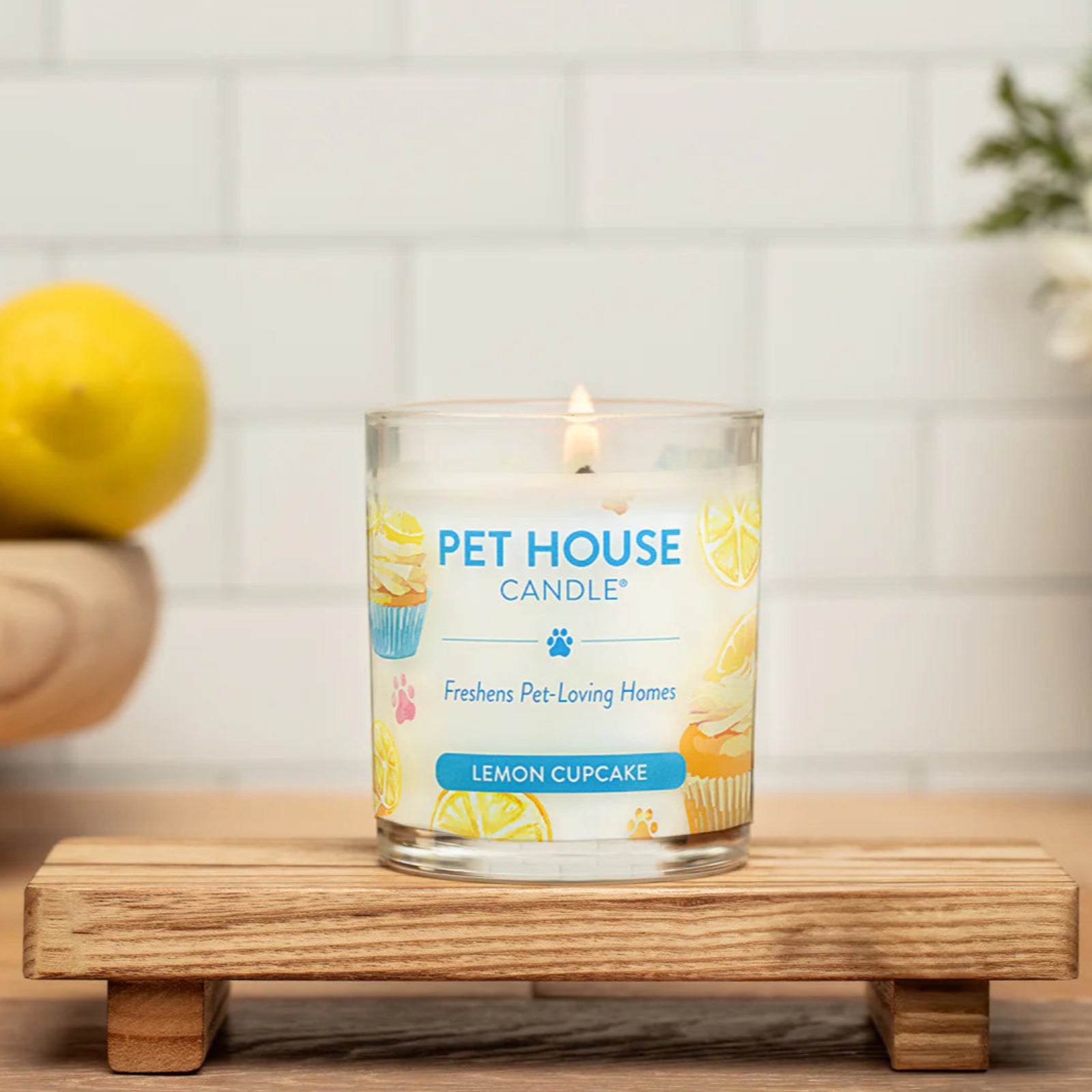 Pet House Candle in Lemon Cupcake flavor on a wooden stand with lemons and flowers in the background.