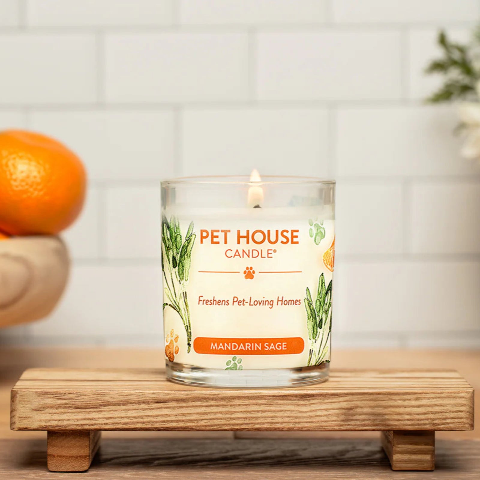 Pet House Candle on a wooden stand with mandarin orange and sage design, surrounded by fruits and flowers.