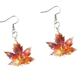 A pair of dangling earrings with silver fishhook wires and leaf-shaped charms in an autumn gradient of orange, red, and yellow on a light cream background.