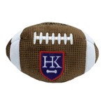A plush football-shaped pet toy in brown and white, featuring a blue and red shield patch with the letters 'HK' and a small white bone.