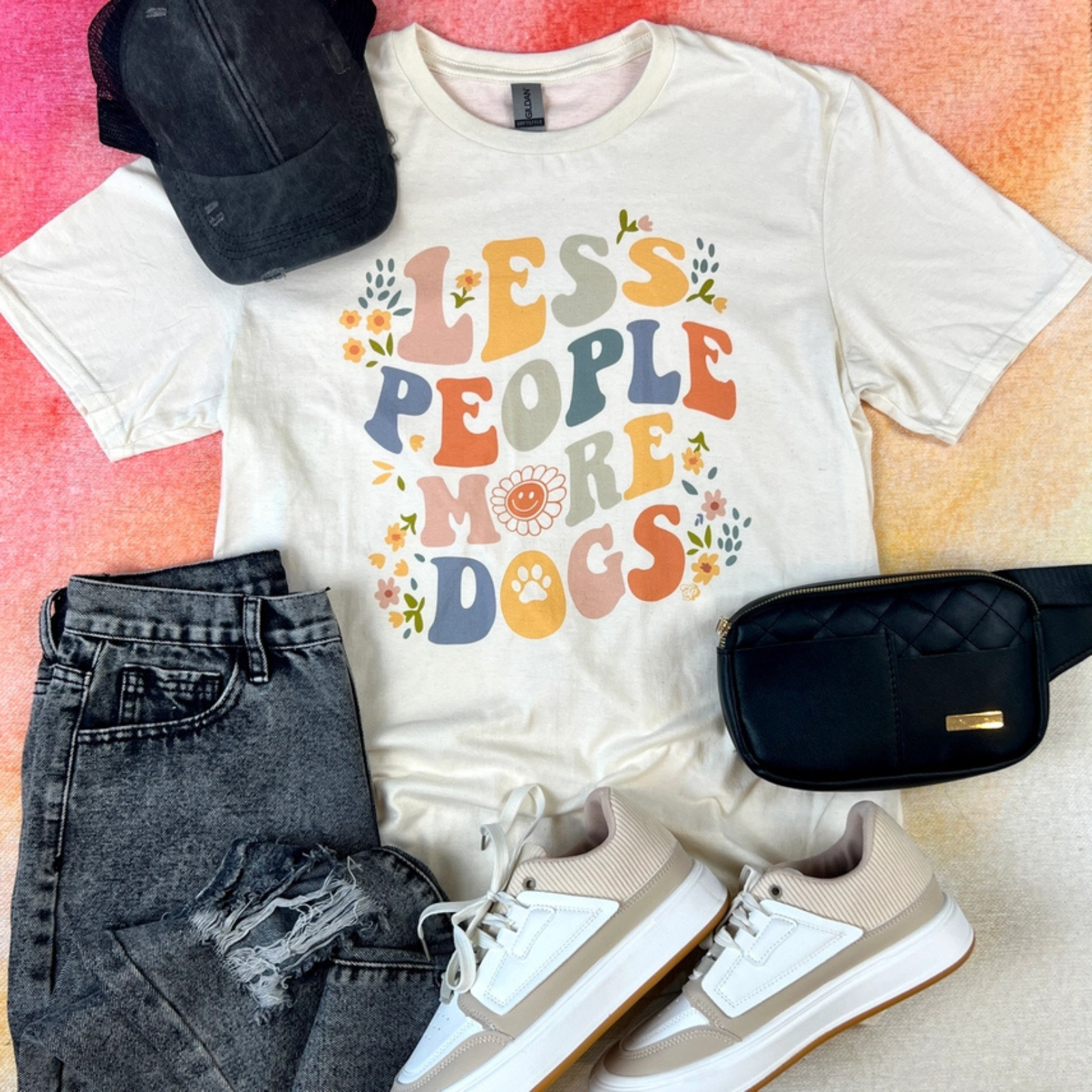 A styled flat-lay outfit featuring a cream graphic t-shirt with the text 'LESS PEOPLE MORE DOGS' in colorful retro letters, paired with distressed dark gray jeans, a black baseball cap, a black quilted crossbody bag, and beige and white sneakers, all arranged on a textured pink and orange background.